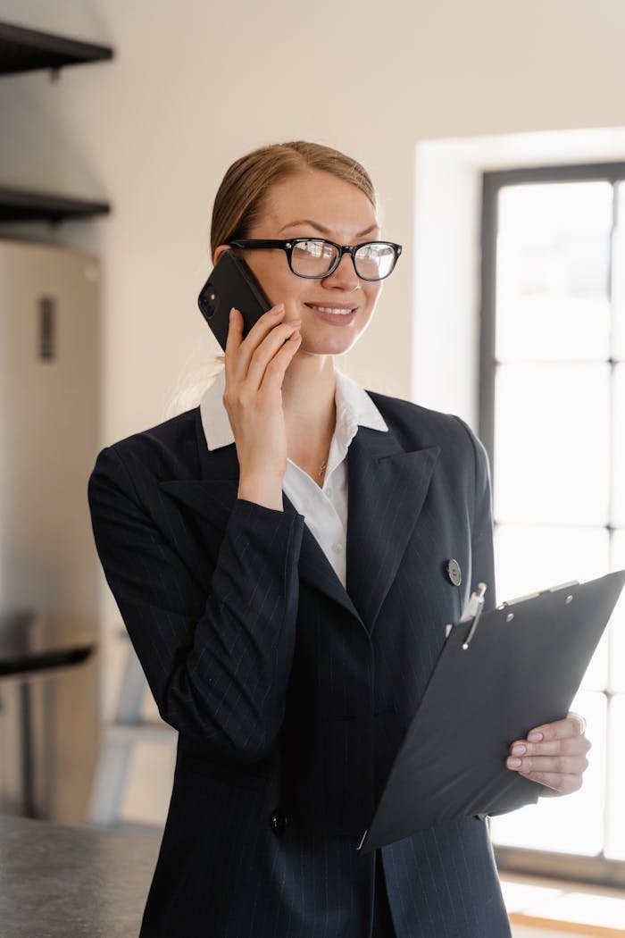 Smiling professional woman in business attire talking on phone holding clipboard in bright office.